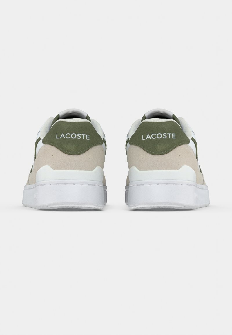Paire de baskets Lacoste blanches et beiges avec des accents verts, vues de dos sur un fond blanc uni.