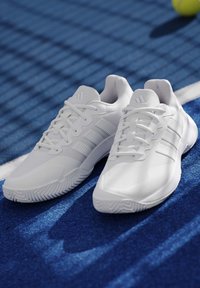 Ζευγάρι λευκών παπουτσιών τένις Adidas που ακουμπούν σε γήπεδο μπλε χρώματος κοντά σε λευκή γραμμή ορίου με μία κίτρινη μπάλα τένις στο φόντο.