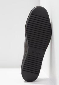 Semelle de chaussure noire avec des rainures horizontales et le logo "Clarks" au centre, positionnée verticalement contre un mur d'angle blanc.