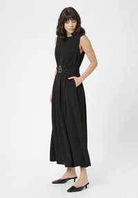 Vrouw in een mouwloze zwarte wijde jumpsuit met riem en zakken, met zwarte lage puntige muiltjes, tegen een effen achtergrond.