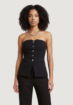 Donna con capelli ricci, indossa un top nero senza spalline con bottoni e pantaloni neri, accessoriata con una collana a catena d'argento.