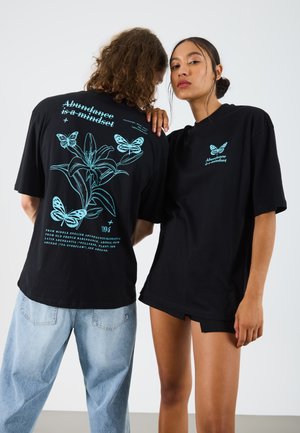 Black oversized T-shirts featuring turquoise butterfly and floral graphics, with text stating "Abundance is a mindset." Casual fit and soft fabric.