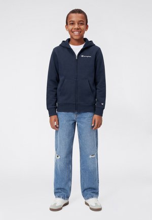 Sudadera con capucha azul marino con cremallera y bolsillo frontal, con logo bordado, combinada con vaqueros desgastados azul claro y zapatos blancos con detalles marrones.