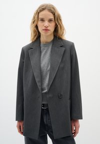 Grå, skræddersyet blazer med en enkelt knaplukning, spidse revers og to forlommer, båret over en grå sweater og mørke jeans.