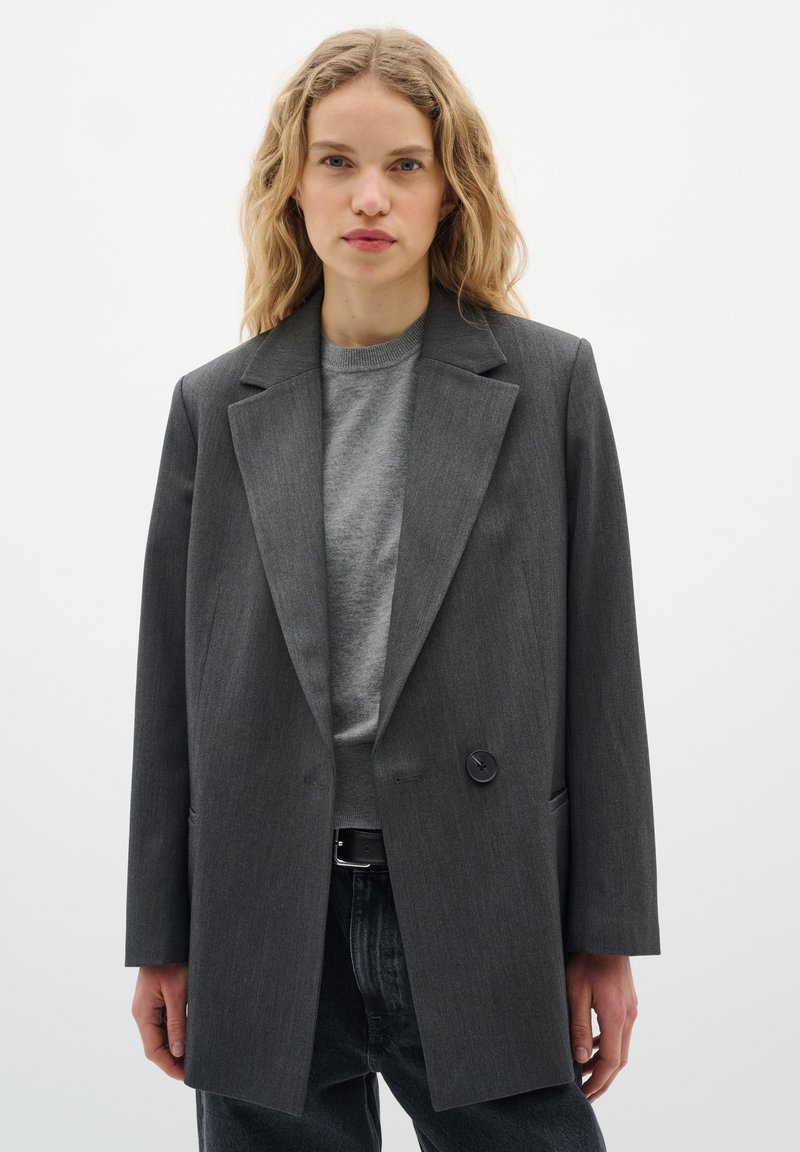 Grå, skræddersyet blazer med en enkelt knaplukning, spidse revers og to forlommer, båret over en grå sweater og mørke jeans.