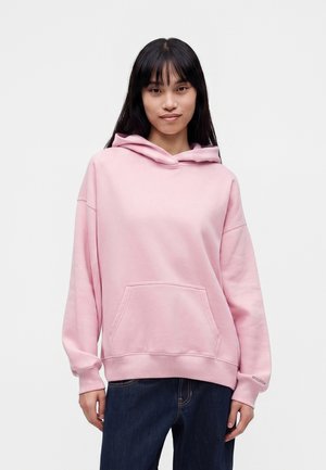 Junge Frau mit langen schwarzen Haaren trägt einen hellrosa Hoodie und dunkelblaue Jeans, steht vor einem einfarbigen hellen Hintergrund.