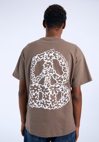 Camiseta de algodón marrón con un gran diseño de símbolo de paz blanco con acentos florales y texto, de manga corta y corte relajado.