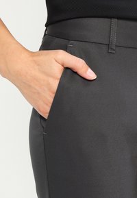 Hand med naturliga naglar som vilar i framfickan på mörkgrå skräddarsydda byxor, bärandes en svart prickig skjorta.