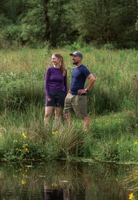 Zwei Personen stehen an einem Teich, der von hohem Gras umgeben ist. Eine trägt ein lila Langarmshirt und schwarze Shorts; die andere trägt ein blaues Shirt und beige Shorts.