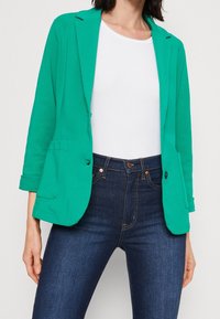 Blazer teal en matériau extensible, avec un col cranté, deux poches avant, des manches retroussées et une coupe décontractée, porté sur un top blanc.