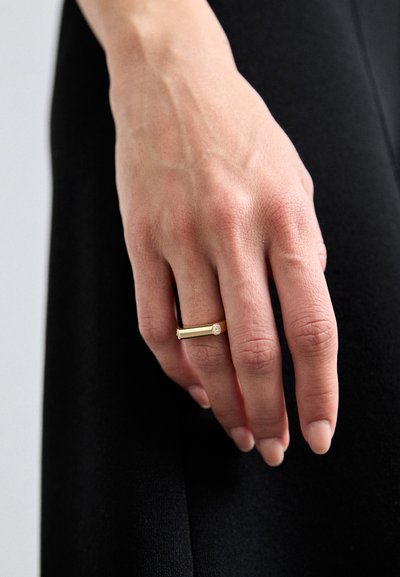 Gouden ring met een rechthoekige band, voorzien van een kleine heldere edelsteenaccent. Glad oppervlak, gedragen aan een hand met natuurlijke nagels.
