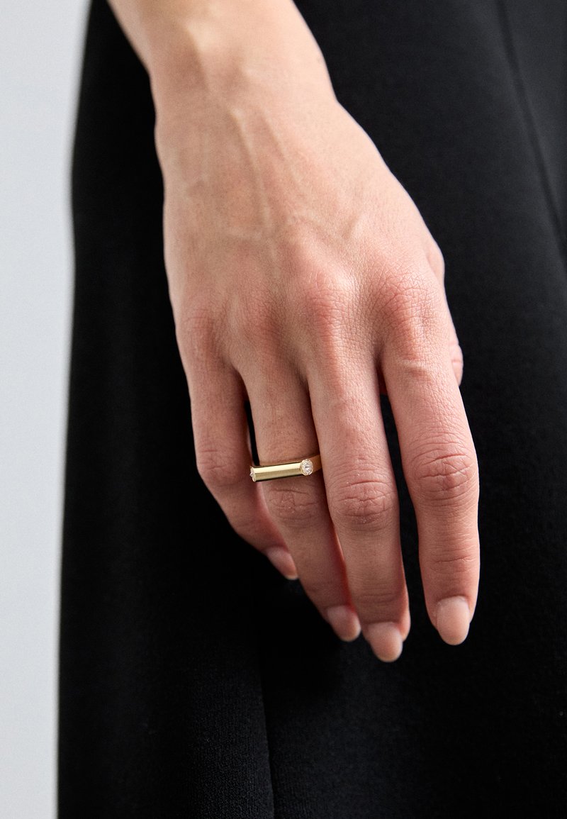 Zelta gredzens ar taisnstūra formu, kam ir mazs caurspīdīgs dārgakmens akcenta elements. Gluda virsma, valkāts uz rokas ar dabīgām nagiem.