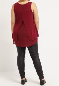 Bordeauxfärgad ärml�ös topp med en delad baksida, lager av tyg, bärs över svarta faux läderleggings med sömdetaljer. Platta svarta skor.