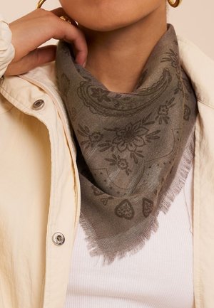Femme portant une écharpe grise à motif floral nouée autour du cou, une veste beige à boutons-pression, un haut blanc côtelé et une boucle d'oreille créole dorée.