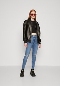 Veste bombardier en cuir noir, crop top noir, jean déchiré bleu clair, bottes chunky noires, lunettes de soleil teintées rouges, texture lisse.