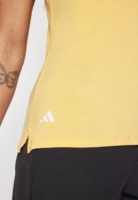 Haut de sport jaune sans manches avec une texture lisse, présentant un logo Adidas blanc et des fentes latérales près de l'ourlet.