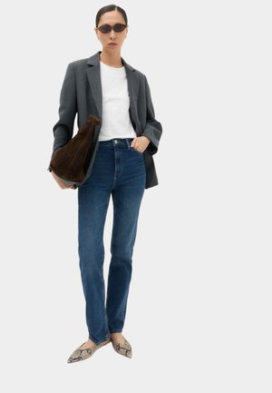 Kvinde iført solbriller, grå blazer, hvid skjorte, blå jeans, holder en brun ruskindstaske og iført flade sko med slangeskindsprint og spids tå.
