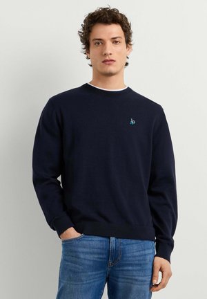 Junger Mann mit lockigem Haar, trägt einen dunkelblauen Pullover mit Rundhalsausschnitt und blaue Jeans, steht mit einer Hand in der Tasche.