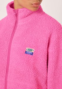 Flis jakna svetlo roza barve z visokim ovratnikom in zadrgo. Na njej je etiketa z napisom "La Maison AMV SUPER VINTAGE" v kontrastnih barvah.