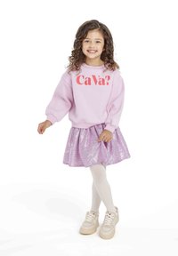 Sweatshirt rose clair avec le texte "Ca Va ?", associé à une jupe en sequins lavande brillante et des collants blancs, complété par des baskets beiges.
