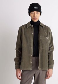 Olijfgroene corduroy overhemd met een knoopsluiting, patch pocket en een getextureerde afwerking, gedragen over een zwarte coltrui. Zwarte beanie op.