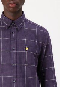 Purple checkered shirt with white grid pattern, button-down collar, and golden logo accent on chest pocket. Featuring dark buttons and textured fabric.