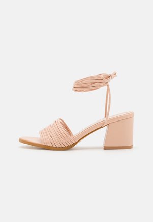 Sandale à talon bloc rose nude avec lanière avant plissée et fine lanière de cheville nouée en un nœud, sur fond blanc.