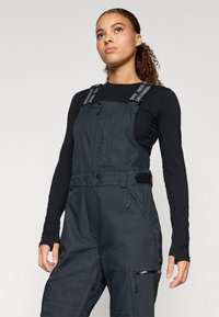 Fekete vászonoverall állítható pántokkal, cipzáras záródással és több zsebbel. Simább, tartós anyagból készült, laza szabású.