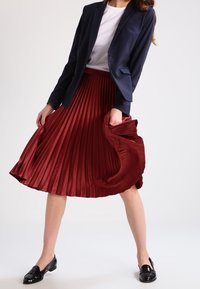 Roter, plissierter Rock mit glatter Satin-Oberfläche, kombiniert mit einem Navy-Blazer und einem weißen Hemd. Schwarze Loafers runden das Outfit ab.