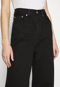 Vaqueros negros de denim de cintura alta con piernas anchas, que cuentan con bolsillos delanteros y detalles de costura visibles, fabricados en tejido de algodón texturizado.