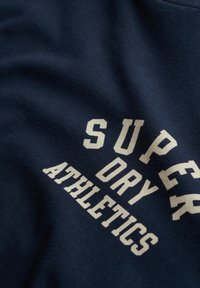 Camiseta de algodón azul marino con texto en color crema que dice "SUPER DRY ATHLETICS", con un acabado texturizado y un diseño casual.