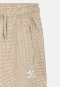 Μπεζ υφασμάτινη φόρμα με ελαστική μέση, πλαϊνές τσέπες και λευκό λογότυπο adidas στην αριστερή πλευρά.