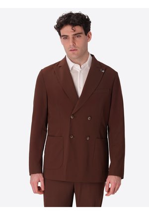 Uomo che indossa una giacca a doppiopetto marrone sopra una camicia bianca con colletto, in piedi contro uno sfondo semplice.