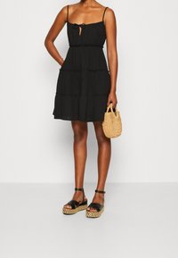 Svart lagerklänning med spagettiband, som har en knytning framtill. Kombinerad med svarta plattformssandaler och en vävd halmhandväska.