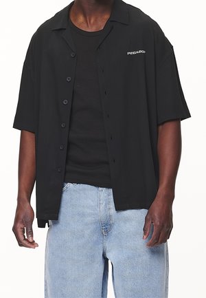 Schwarz kurzärmliges Hemd mit lockerer Passform, Knopfleiste und Logodetail auf der Brust, kombiniert mit hellblauen Jeans.