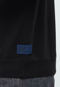 Primer plano del dobladillo de sudadera negra con etiqueta azul que dice "Dedicado a la Perfección del Denim", usada sobre jeans de mezclilla grises.
