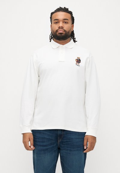 Polo blanco de manga larga con cuello, que presenta un logo de oso bordado en el pecho y puños acanalados. Combinado con vaqueros azul oscuro.