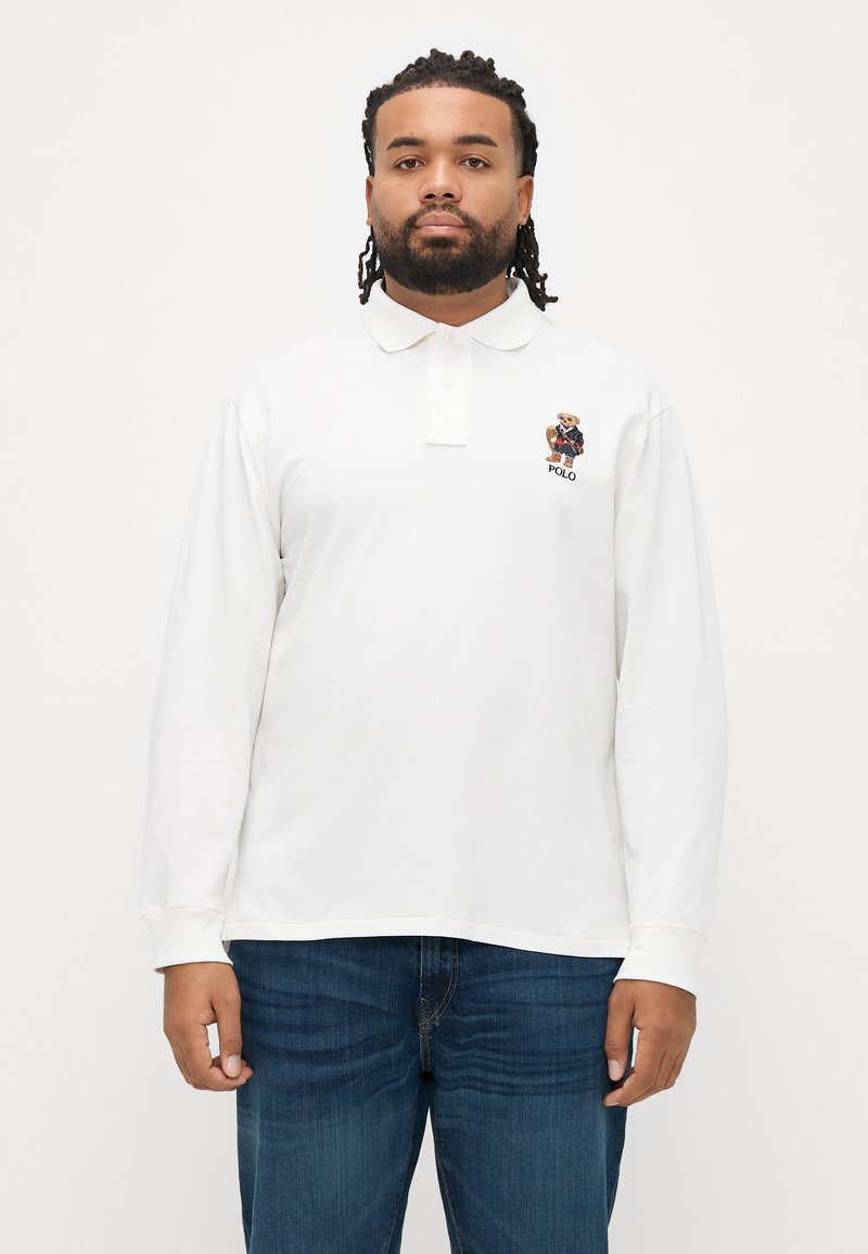 Λευκό πόλο πουκάμισο με μακριά μανίκια και γιακά, με κεντητό λογότυπο αρκούδας στο στήθος και ριμπ μανσέτες. Φορεμένο με σκούρα μπλε τζιν.