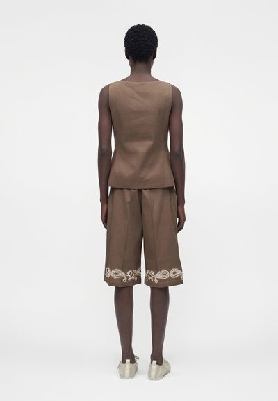 Άτομο με πλάτη, φοράει καφέ αμάνικη μπλούζα και φαρδιές σορτς με λευκό κέντημα paisley κοντά στο τελείωμα, λευκά αθλητικά παπούτσια.