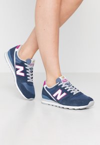 Marineblå sneakers med rosa accenter och vit logga, som har en blandning av mesh och mocka material, grå snörning och en vit gummisula.