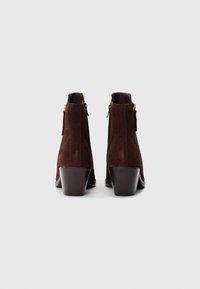 Bottines en daim marron à talon moyen, avec une fermeture éclair latérale et des accents en métal doré. Texture lisse et design classique.