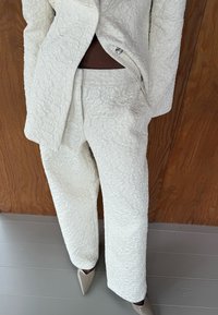 Costume en tissu blanc texturé comprenant un blazer et un pantalon décontracté, avec un motif en relief et des fermetures à boutons argentés, associé à des talons de couleur claire.