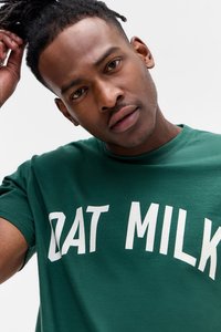 Green cotton t-shirt featuring bold white lettering spelling "OAT MILK." Crew neckline and short sleeves, smooth texture.