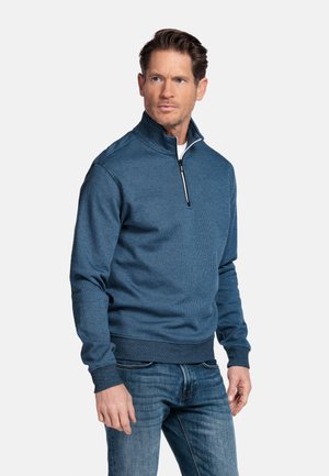 Man draagt een blauw pullover met korte ritssluiting en jeans, staat en kijkt opzij tegen een effen lichte achtergrond.