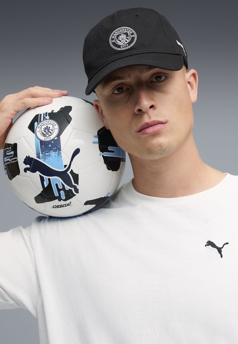 Homme portant une casquette noire de Manchester City et un T-shirt blanc Puma, tenant un ballon de football blanc Puma et Manchester City sur l'épaule.