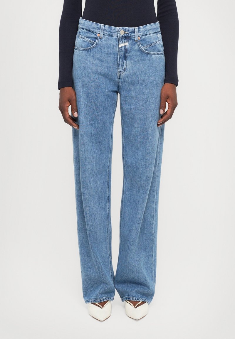 Personne portant un jean bleu clair à jambes droites, un haut noir à manches longues et des chaussures blanches à bouts pointus, debout devant un fond uni.