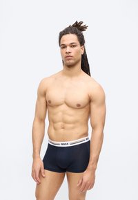 Homme musclé aux longues dreadlocks se tient devant un fond blanc, portant un boxer noir BOSS, face à la caméra avec une expression neutre.