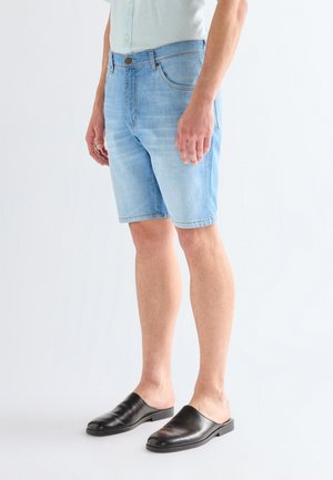 Mann trägt hellblaue Jeansshorts, ein helles Hemd und schwarze Slip-On-Schuhe und steht vor einem weißen Hintergrund.