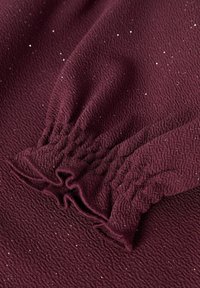 Tissu texturé bourgogne avec un détail smocké froncé et des accents de brillance subtils. La surface est légèrement froissée, ajoutant de la profondeur.