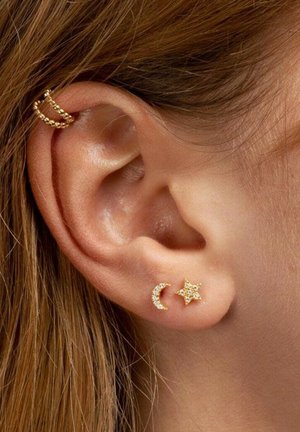 Orecchini in tonalità oro con design a mezzaluna e stella, adornati con gemme trasparenti, su uno sfondo in tonalità della pelle.